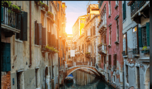 Narrow Venice canal at sunset with a stone pedestrian bridge, historic palaces and a moored boat