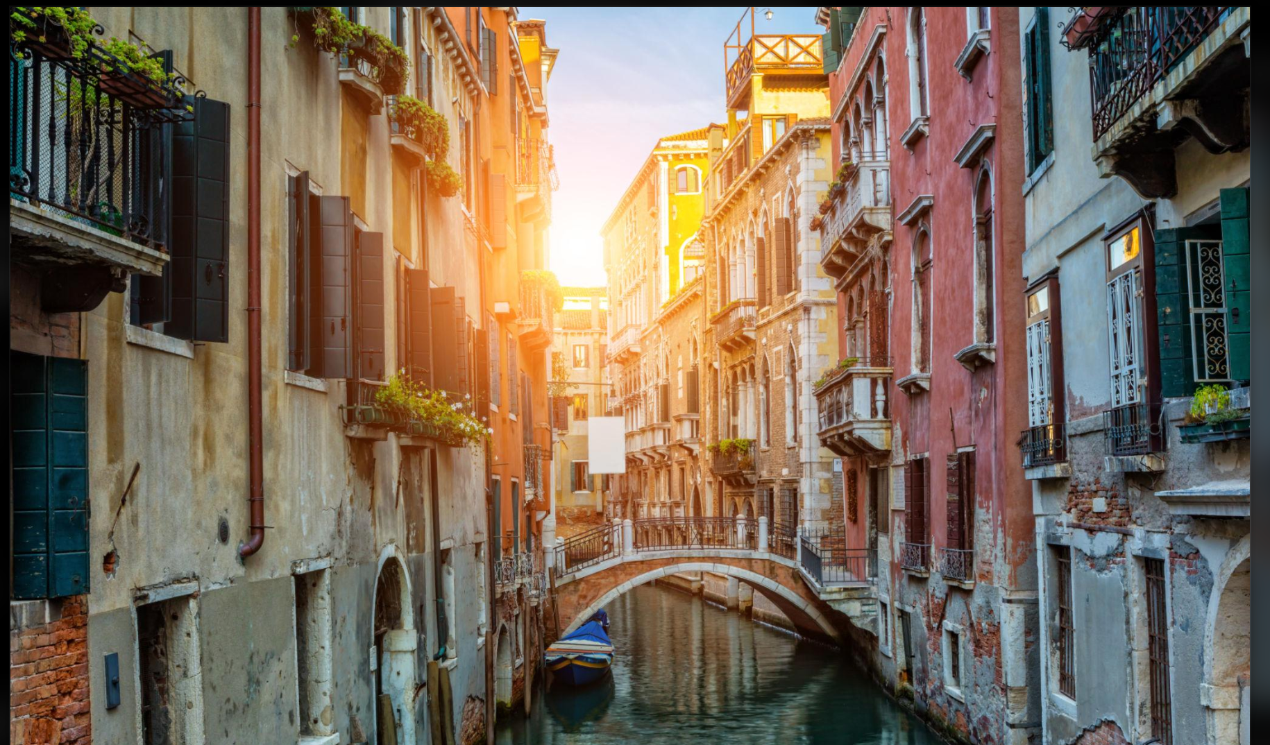Narrow Venice canal at sunset with a stone pedestrian bridge, historic palaces and a moored boat